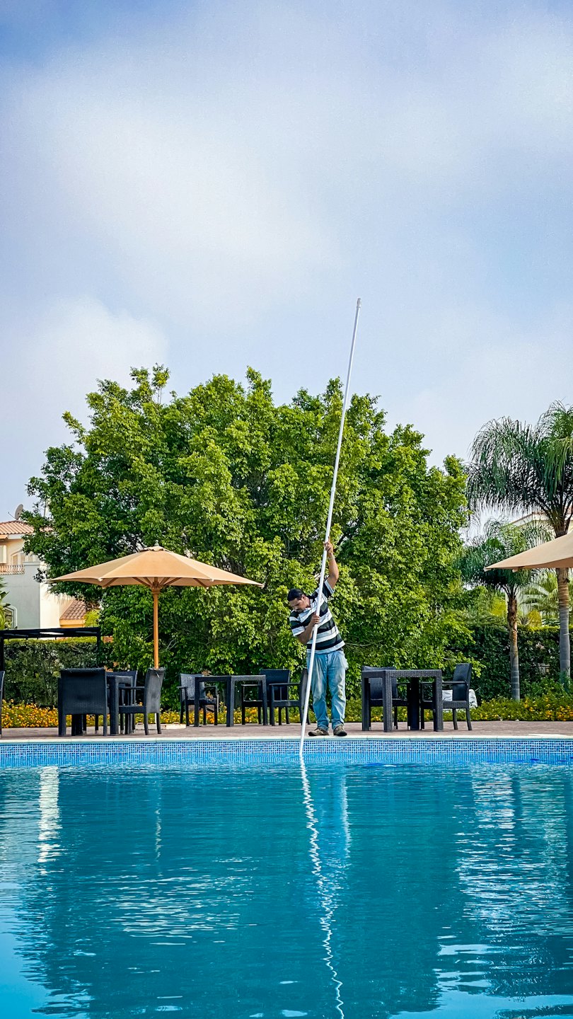 Pool worker cleans large swimming pool with long pole.